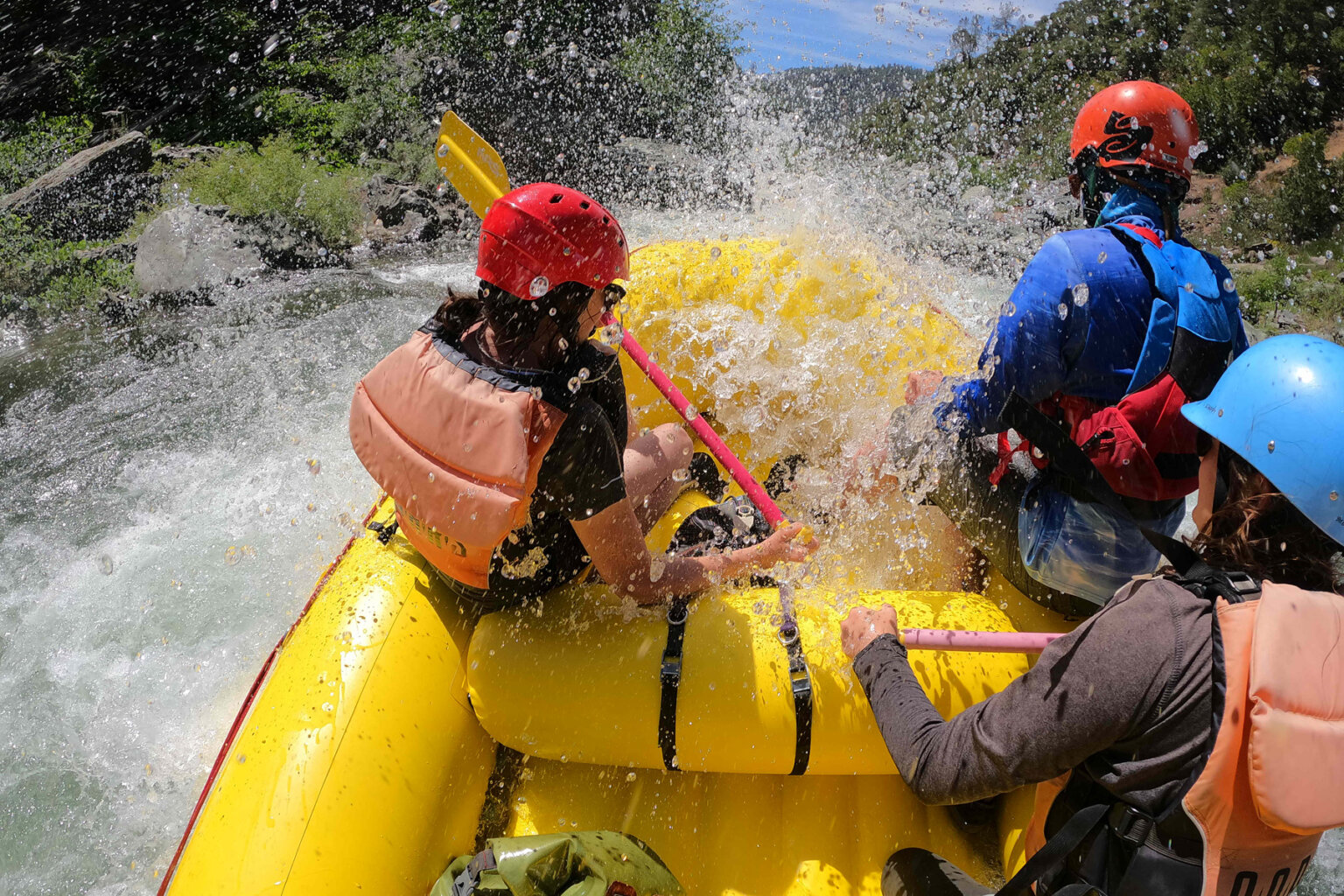 View from behind of three paddlers in the front sections of a yellow raft as they get soaked by whitewater spray.