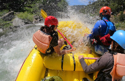 View from behind of three paddlers in the front sections of a yellow raft as they get soaked by whitewater spray.