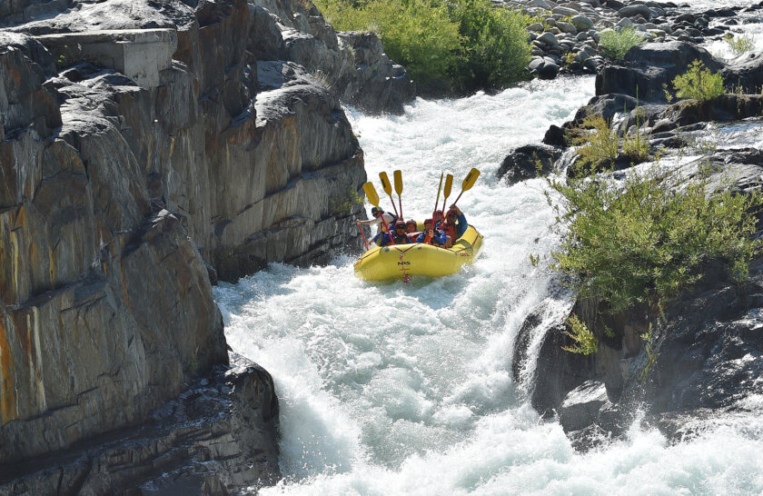 A group of paddlers get down in a yellow raft with their paddles held straight in air as a guide paddles as they enter frothy Tunnel Chute rapid.