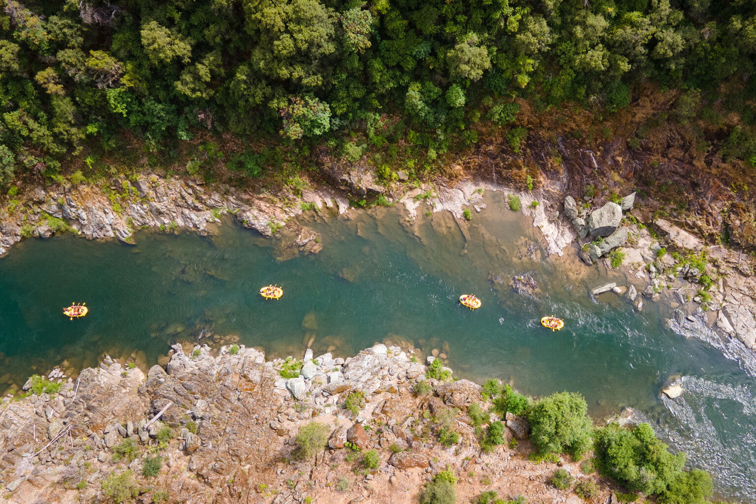 Aerial view of four yellow rafts as they make their way down the Middle Fork American River surrounded by rocky tree-studded shores.