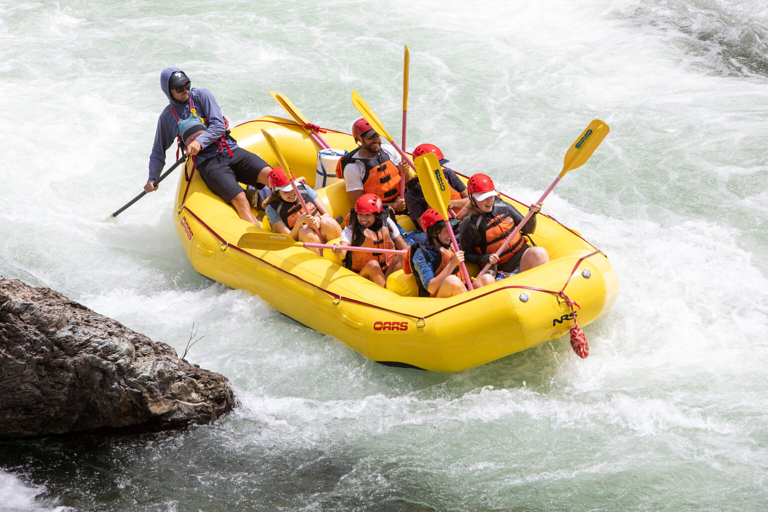 A yellow raft steered by a guide goes through frothy whitewater as smiling guests get down in the boat.
