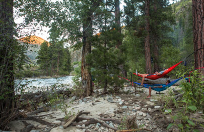 Two guests lay back in hammocks amongst the trees on the shore of the Middle Fork of the Salmon River in Idaho.
