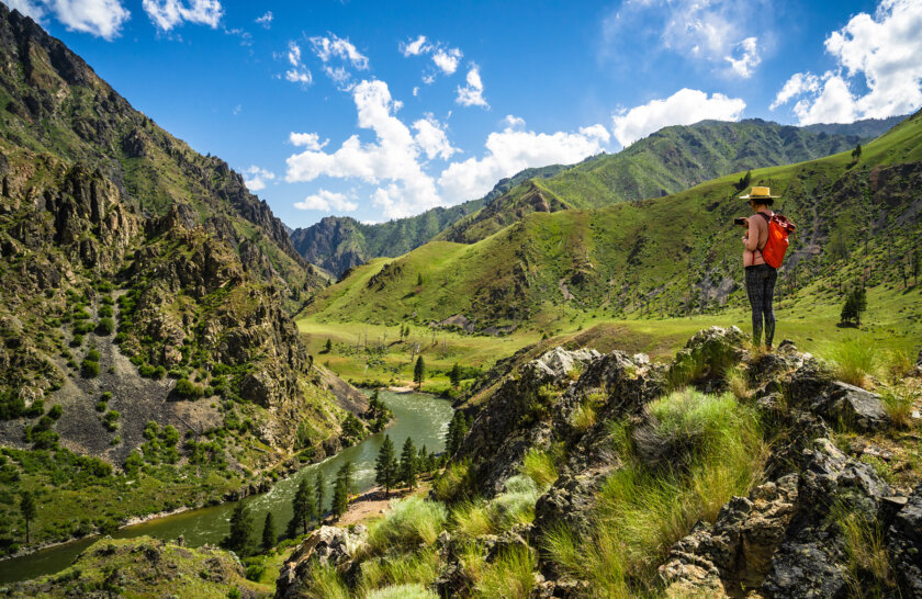 A hiker stands on a boulder high above the Salmon River and densely lush hillsides and mountains rise in the distance.