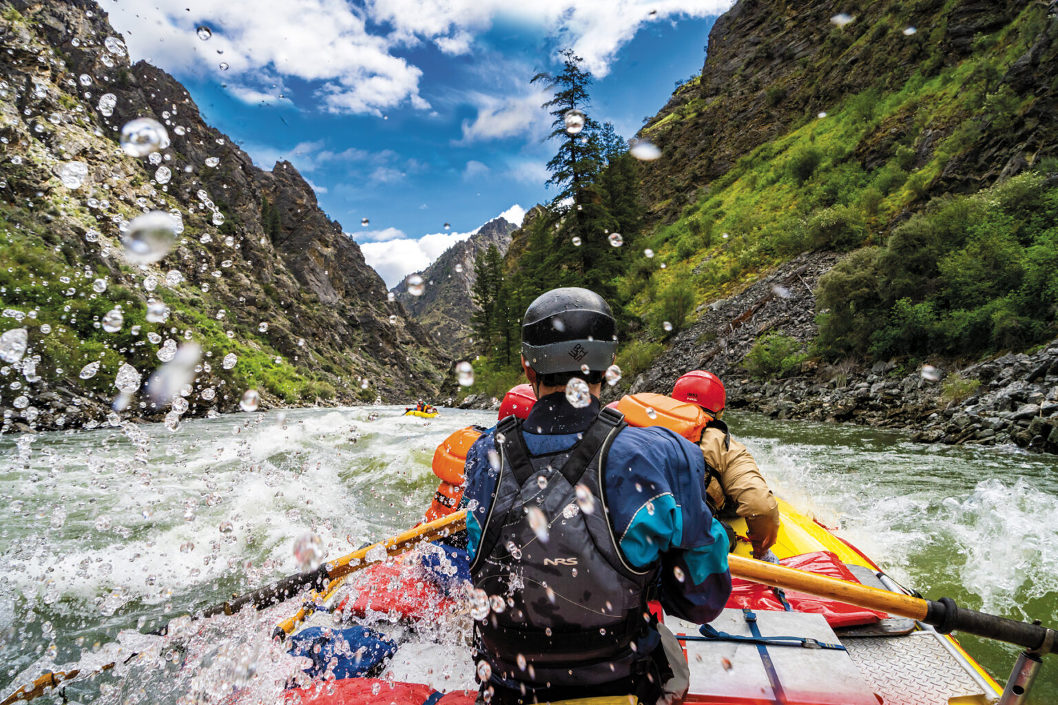 View from behind of a guide rowing their raft through whitewater on the Middle Fork Salmon River and large water spray surrounds them.