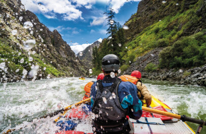 View from behind of a guide rowing their raft through whitewater on the Middle Fork Salmon River and large water spray surrounds them.