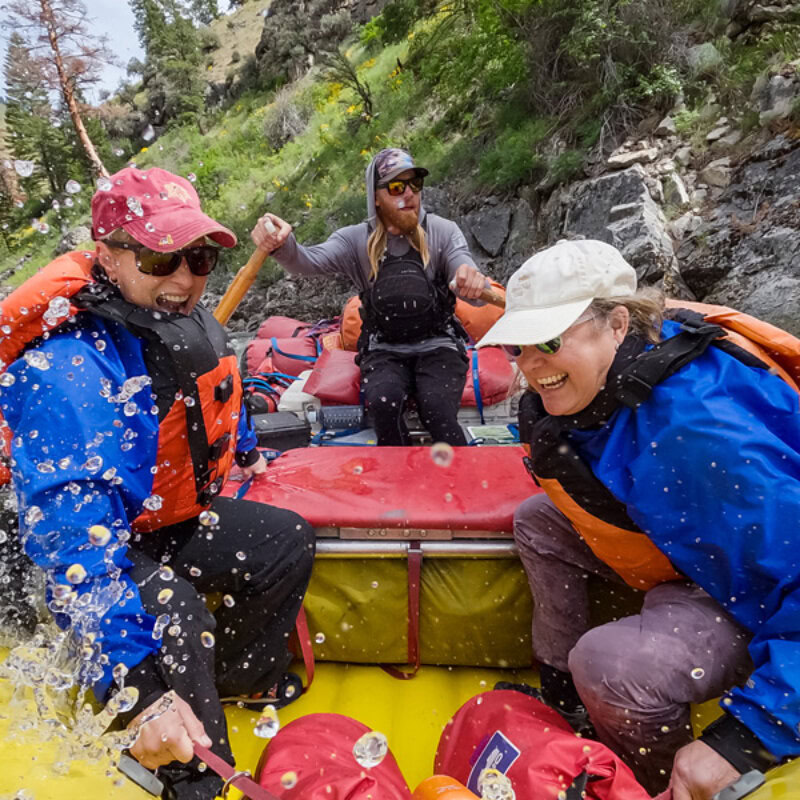 Two laughing women hold on as they get splashed going through a rapid wile their guide rows behind them on the Middle Fork of the Salmon River.