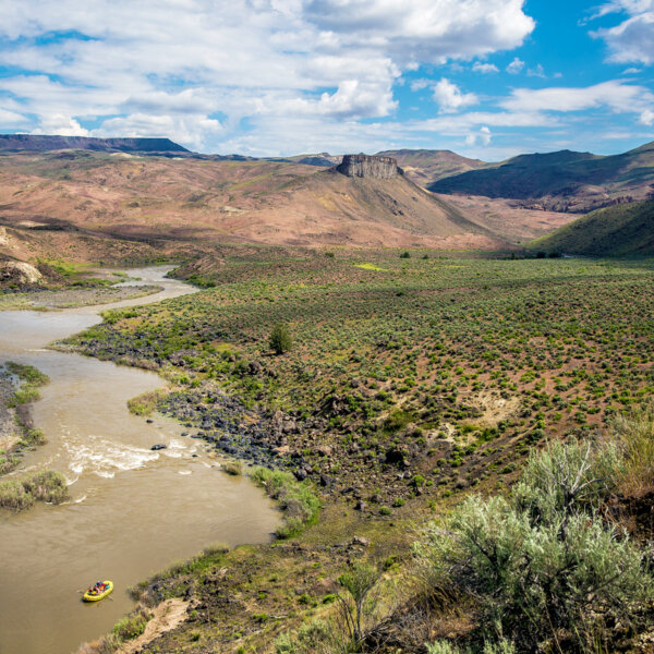 A vast stretch of land with mountains in the background and a yellow raft rowing down the Owyhee River.