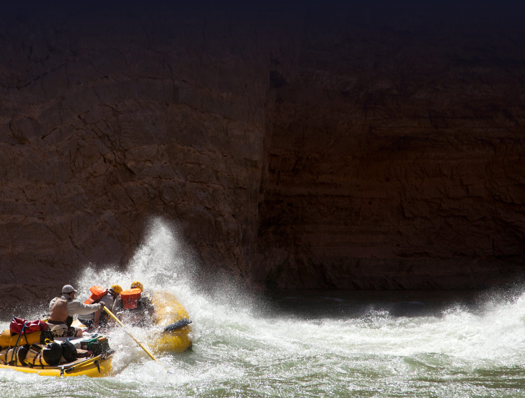 A yellow raft plows through a wave on the river with canyon walls rising above.