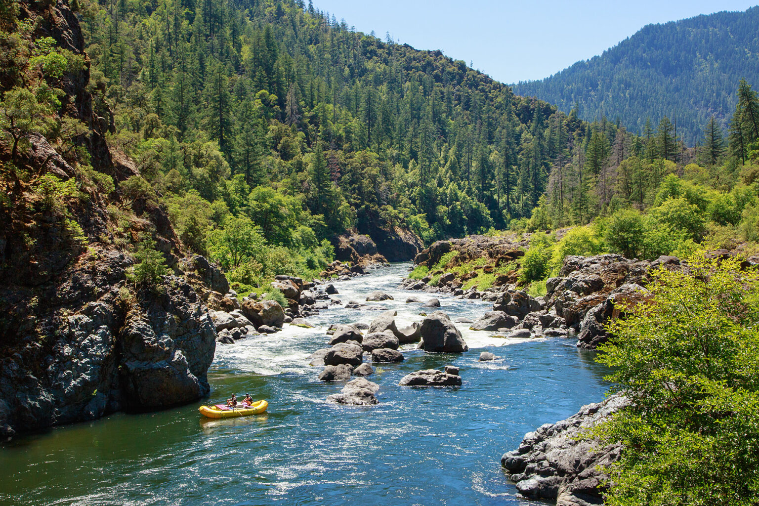 Scenic photo of a yellow raft entering Blossom Bar Rapid in a lush, forested valley on the Rogue River