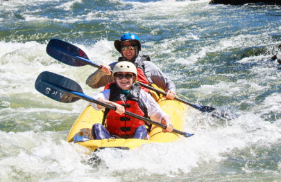 Helmeted duo smile for the camera as they paddle their yellow inflatable kayak through light rapids on the Rogue River