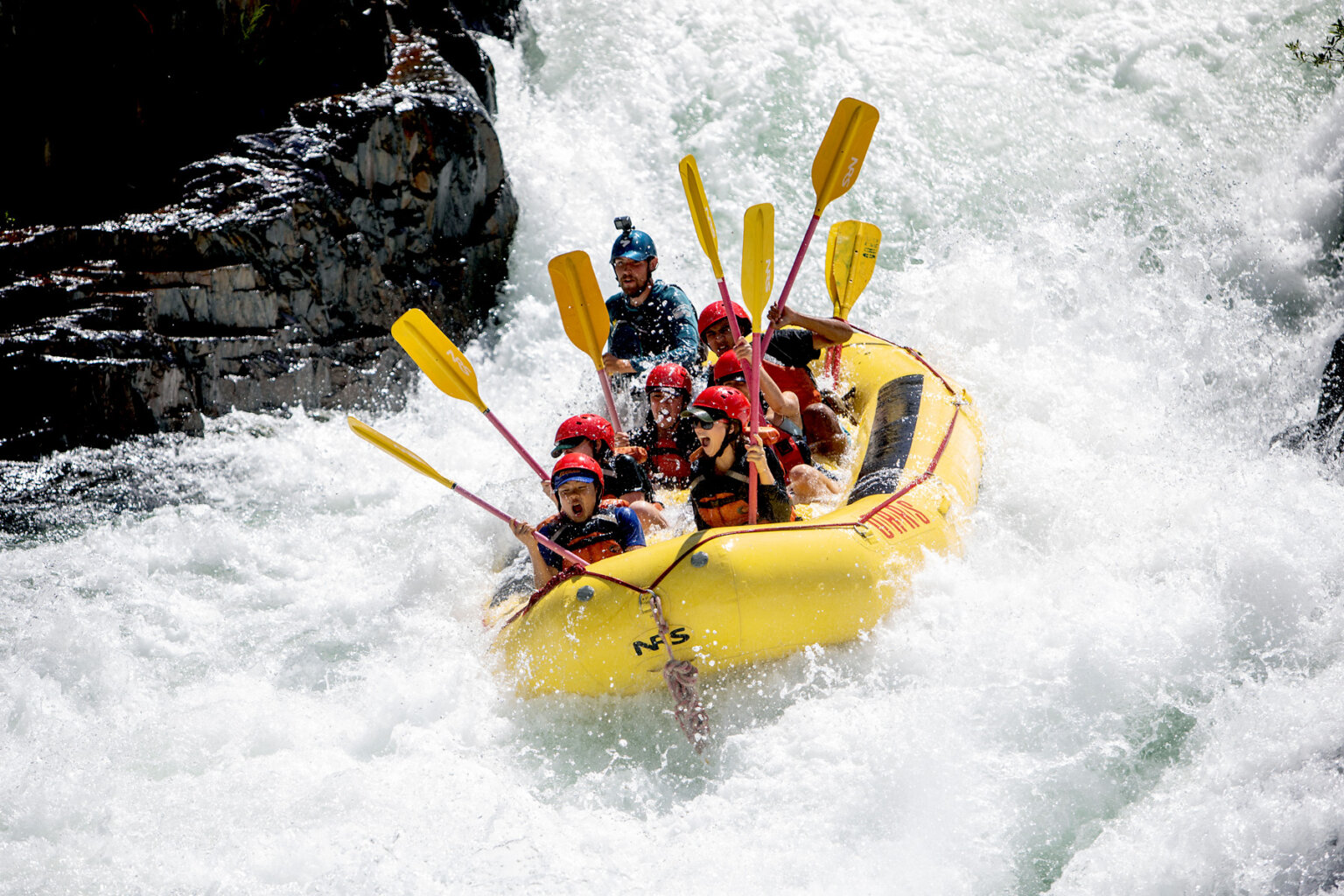 A group of paddlers get down in a yellow raft with their paddles held straight in the air as a guide paddles through a monstrous frothy white rapid.