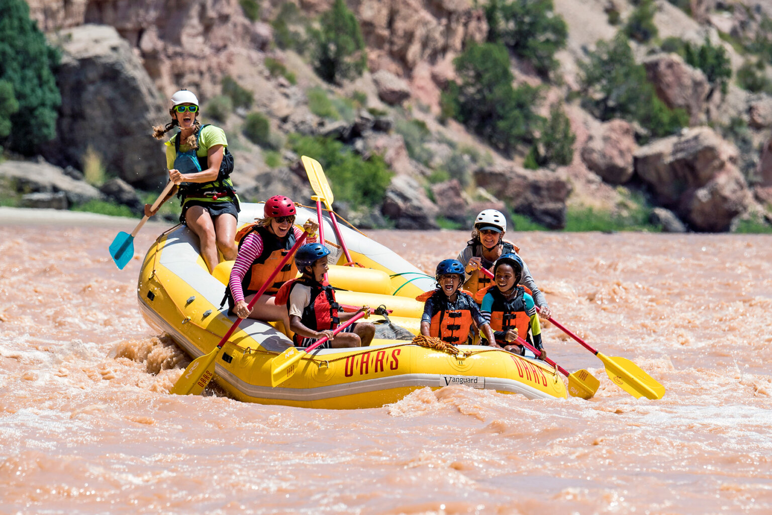 Three ecstatic children in the front of a paddle raft with two adults and a guide behind them as they paddle through a small rapid on the Green River.