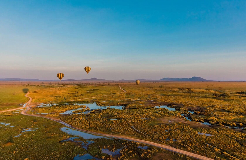 Aerial view of a vast landscape with three striped hot air balloons drifting over greenery and a winding dirt road, under a clear blue sky.