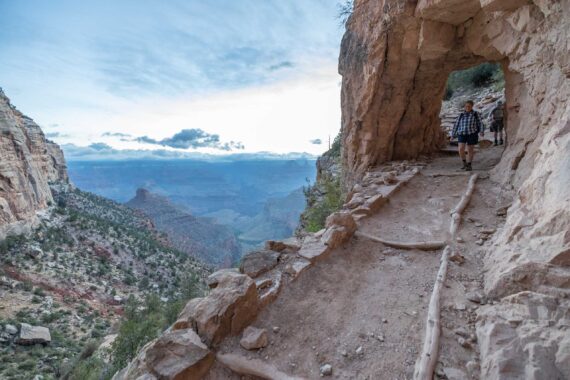 2 people walking through a hole in the rock on a trail next to a cliff.