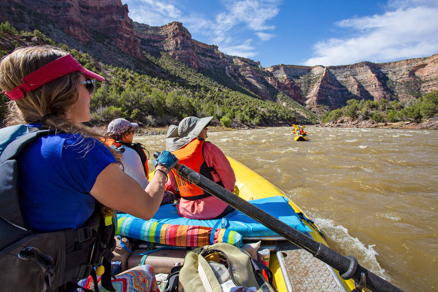 Guests sit in the front of a raft and look up at the view from the Yampa River while a guide rows them downstream.