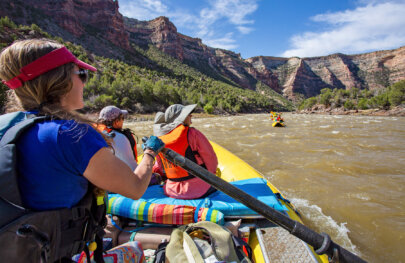 Guests sit in the front of a raft and look up at the view from the Yampa River while a guide rows them downstream.