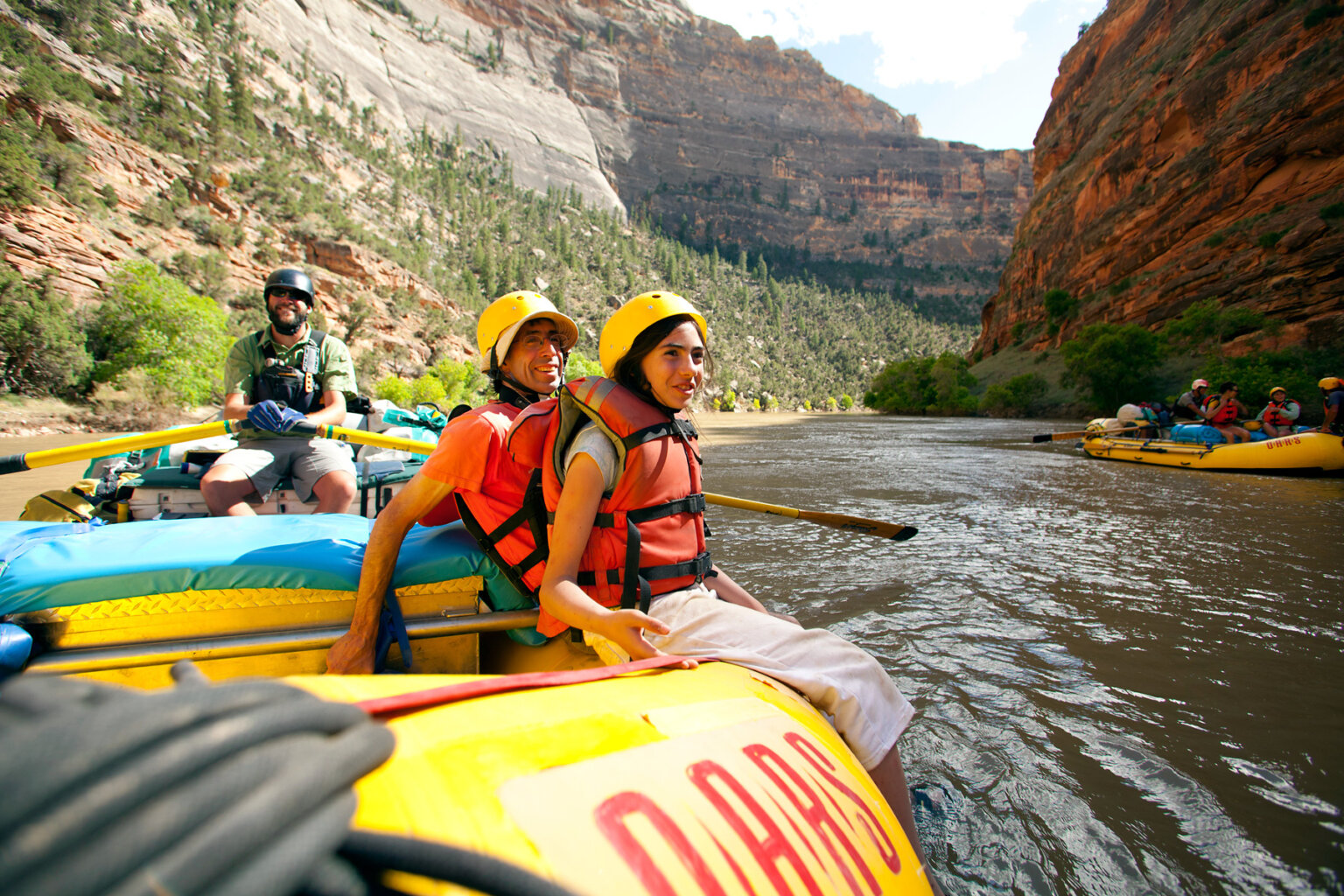 A man and his daughter sit on the tubes of a raft with their feet dangling over the river and smile as they take in the sites while their guide rows them downstream.