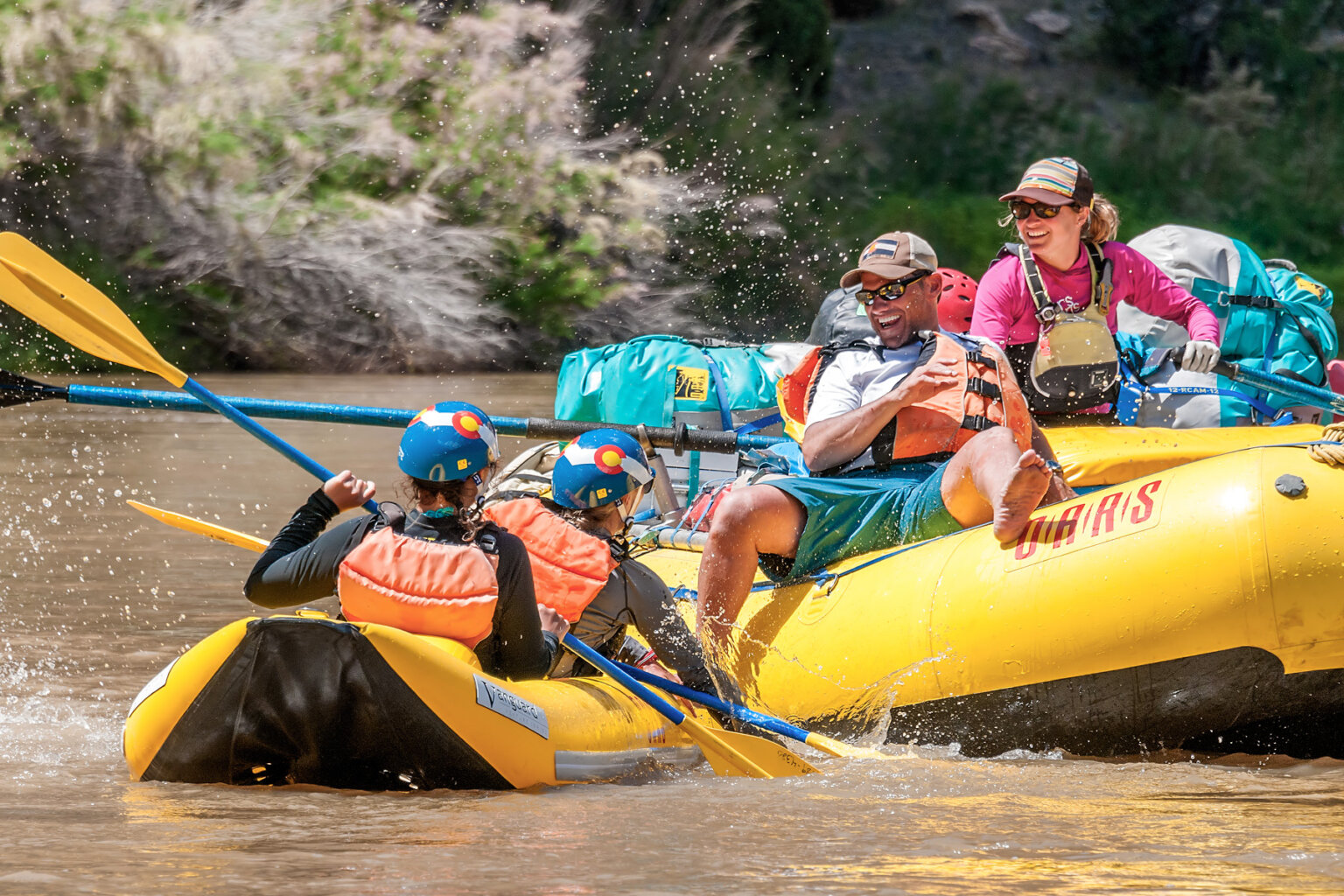Two guests in an inflatable kayak have a water fight with another guest sitting on the tube of a yellow raft, while a smiling guide watches.