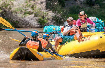 Two guests in an inflatable kayak have a water fight with another guest sitting on the tube of a yellow raft, while a smiling guide watches.