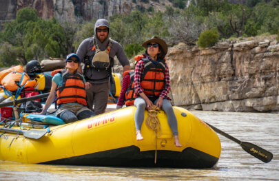Two guests sit at the front of a yellow raft and look up at the scenery while their guide rows them downstream on the Yampa River.